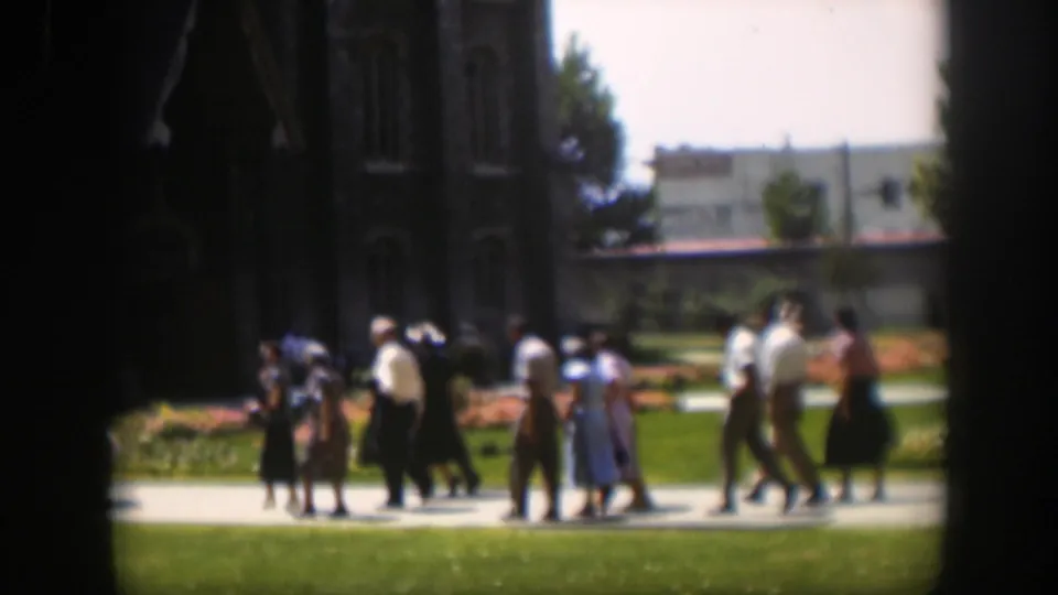 Wyoming 1951 home-movie frame with adults walking together on a path, seen through dark vignetted edges near a large building.