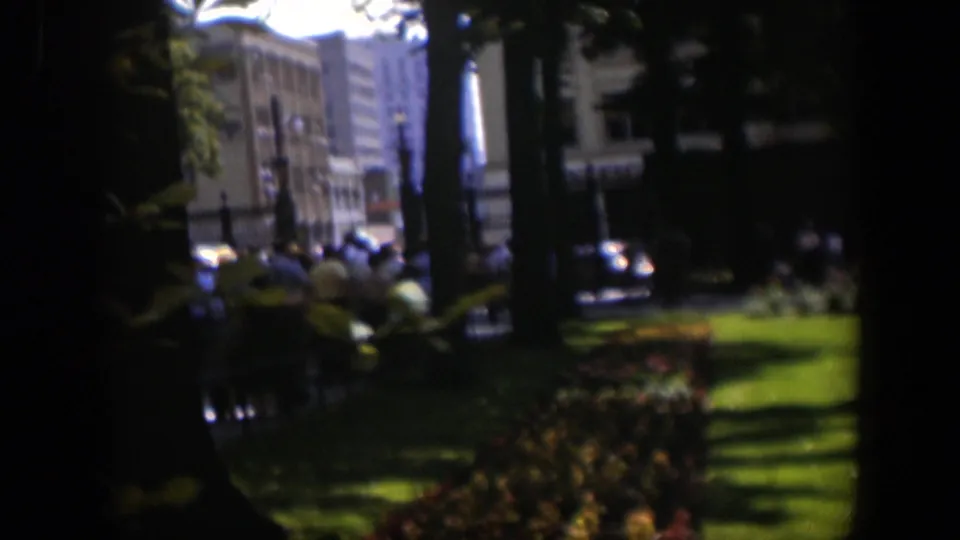 Soft 8mm frame of a 1951 Wyoming churchyard or park edge, with people by the fence and mid-century streetscape details in the background.
