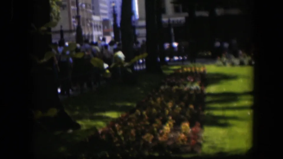 Later 8mm frame from the 1951 Wyoming scene showing a churchyard lawn, flowerbed, fence, people, parked cars, and town buildings.