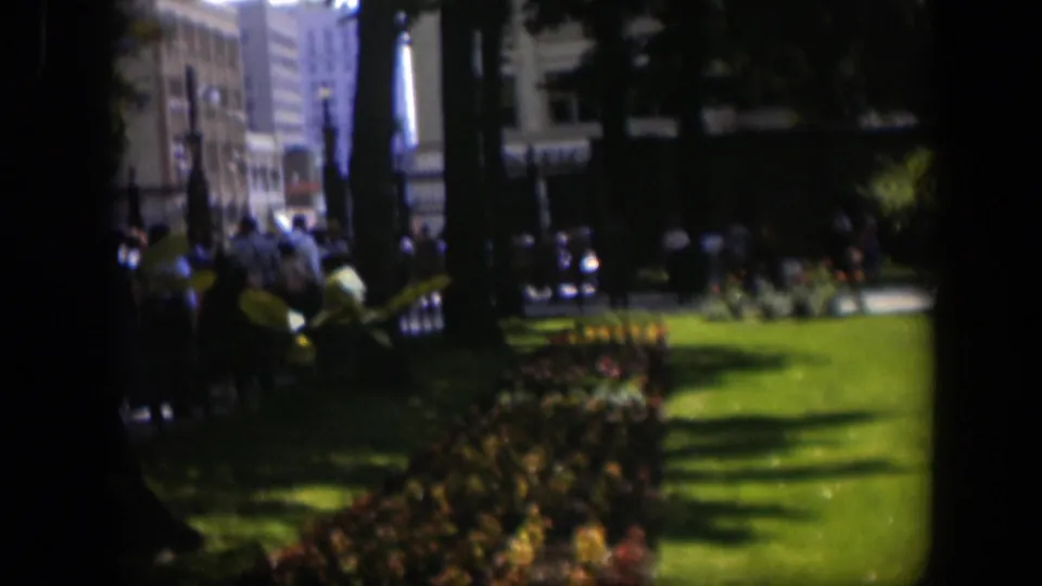 1951 Wyoming 8mm frame showing a green park lawn, flowerbeds, tall trees and distant people walking near a path.
