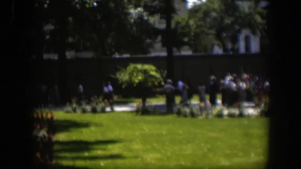 1951 Wyoming home-movie frame with a larger group of distant people near flowerbeds, a green lawn and tall trees.