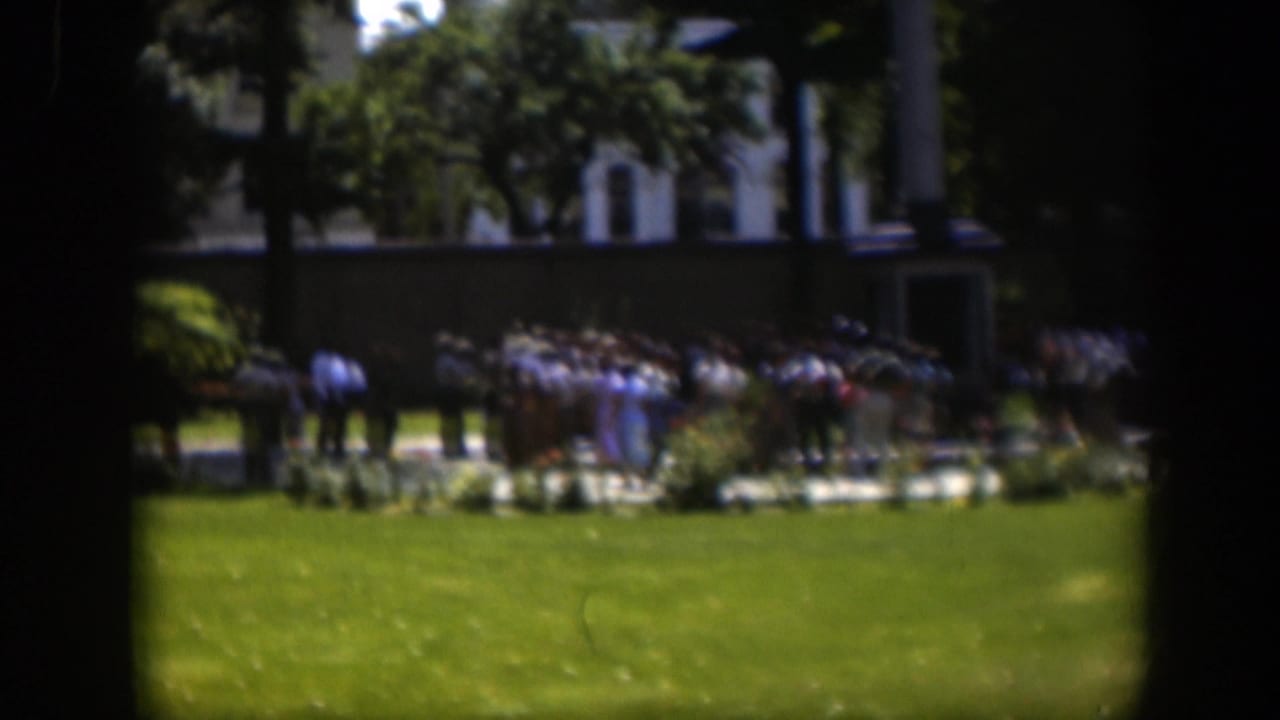 Crowd clustered near plantings and a pale building in a bright park setting.