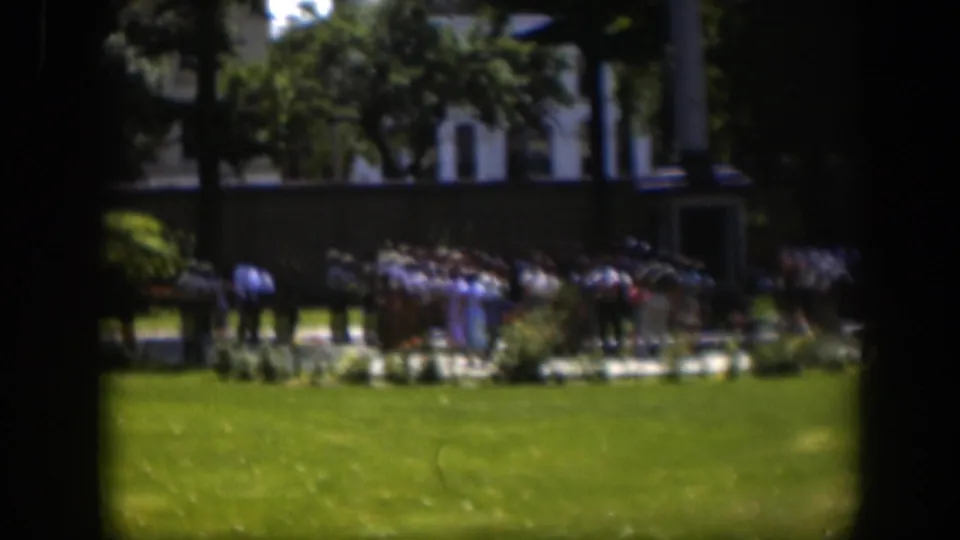 1951 Wyoming home-movie frame with a distant crowd near landscaped beds and trees, framed by dark vertical edges.