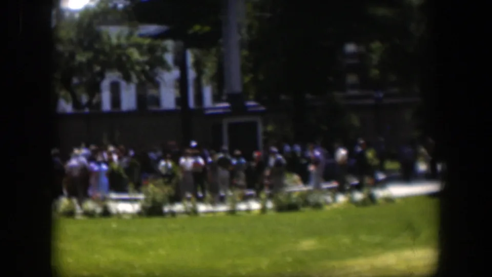 Distant 1951 Wyoming park gathering on 8mm film, with people lined near a walkway and building entrance.