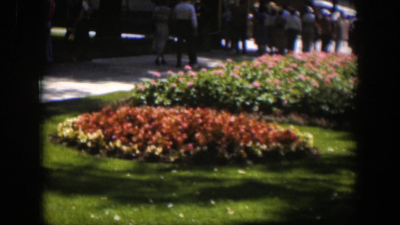 Red and pink flower beds beside a green lawn with distant people walking on a path.