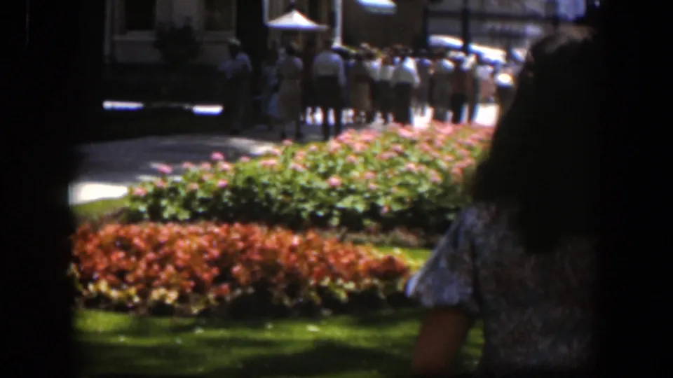 A 1951 Wyoming park view on 8mm film, with flower beds in sunlight and a foreground passerby partly blocking the right edge.