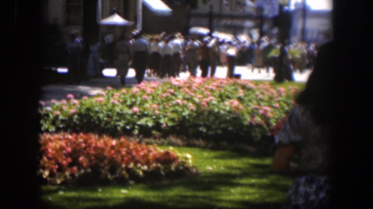 1951 Wyoming park scene with red flowers, green lawn, a woman in a patterned skirt, and crowds walking near buildings.