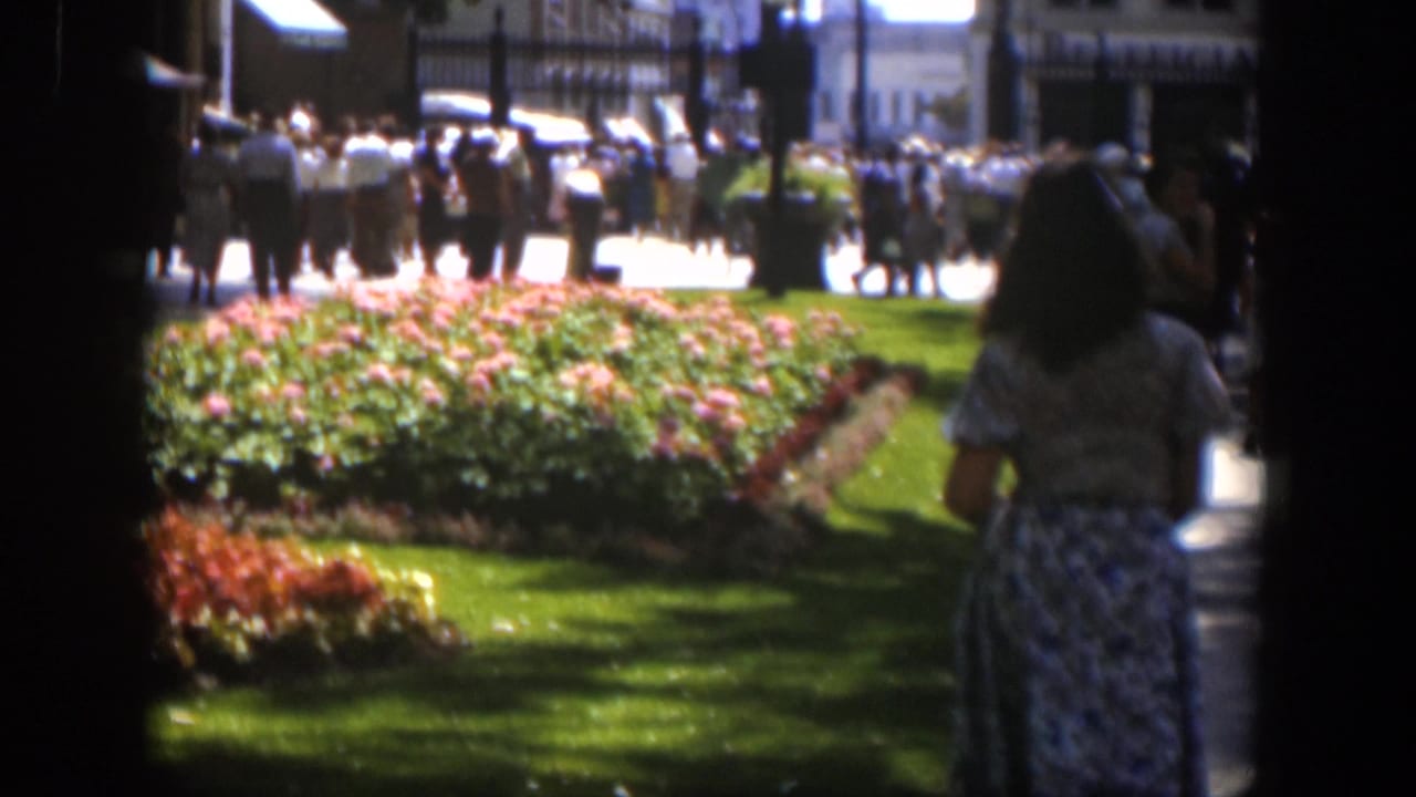 Woman walks at the right edge of a park scene with flowerbeds and a crowd in the background.