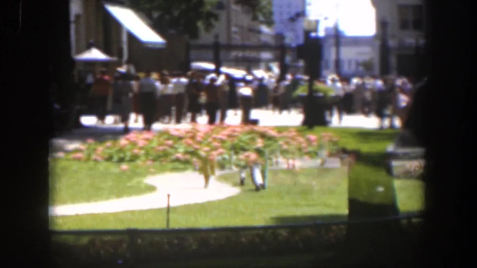 Sunlit Wyoming park view with a curved path, bright flowerbeds, green grass, and mid-century pedestrians in soft 8mm focus.