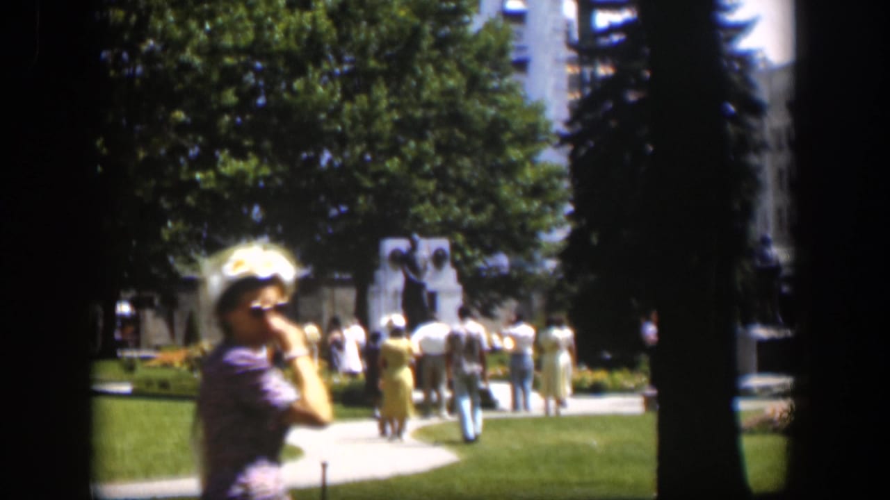Woman in a hat passes near the camera as people gather by a park monument.