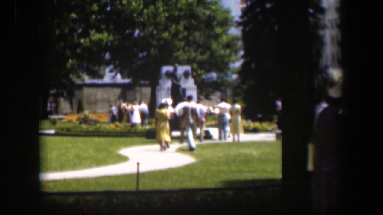 Visitors in 1950s clothing move along a curving path toward a monument in a sunny Wyoming park, framed by dark tree trunks.