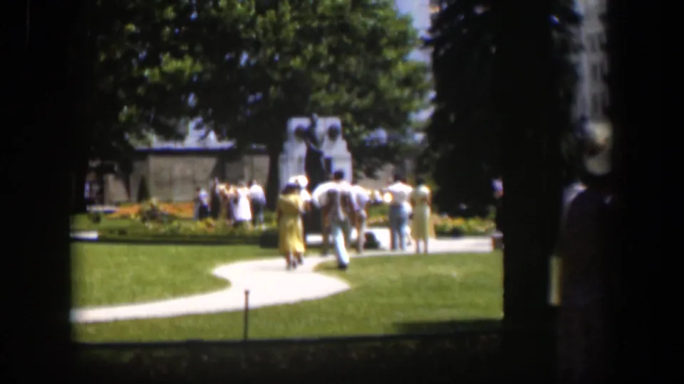 Visitors in 1950s clothing move along a curving path toward a monument in a sunny Wyoming park, framed by dark tree trunks.