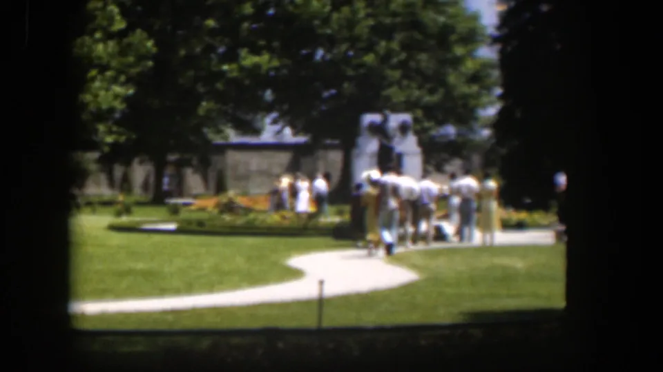 A small group in light summer clothing gathers near a park monument, with grass, trees, and a curved path visible in the 1951 Wyoming footage.
