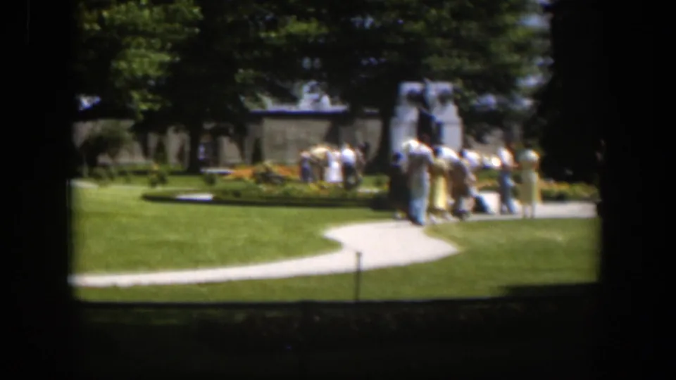 People cluster near a monument in a sunny Wyoming park, with a winding path, broad lawn, trees, and 8mm frame shadows around the scene.