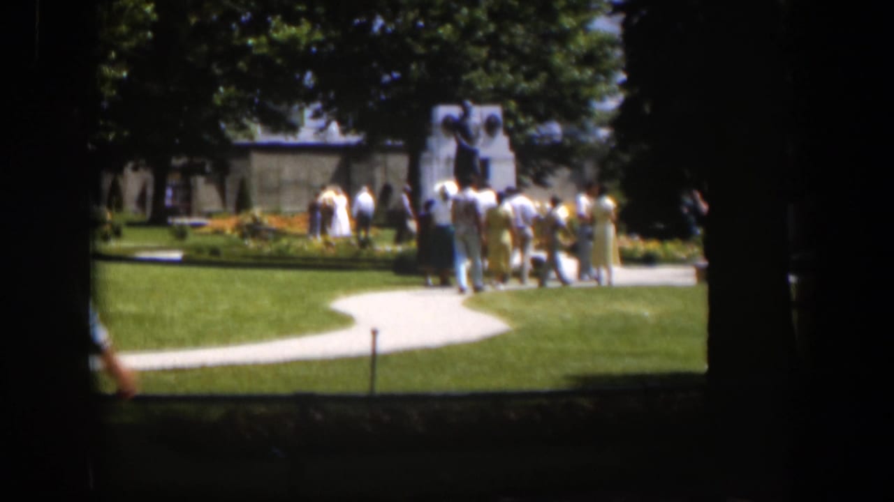 1951 Wyoming 8mm frame showing visitors in a landscaped park with curving paths, trees, flowerbeds, and a monument in the distance.
