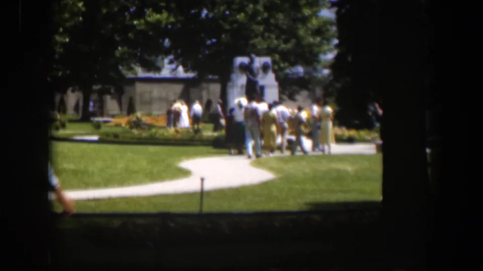 1951 Wyoming 8mm frame showing visitors in a landscaped park with curving paths, trees, flowerbeds, and a monument in the distance.