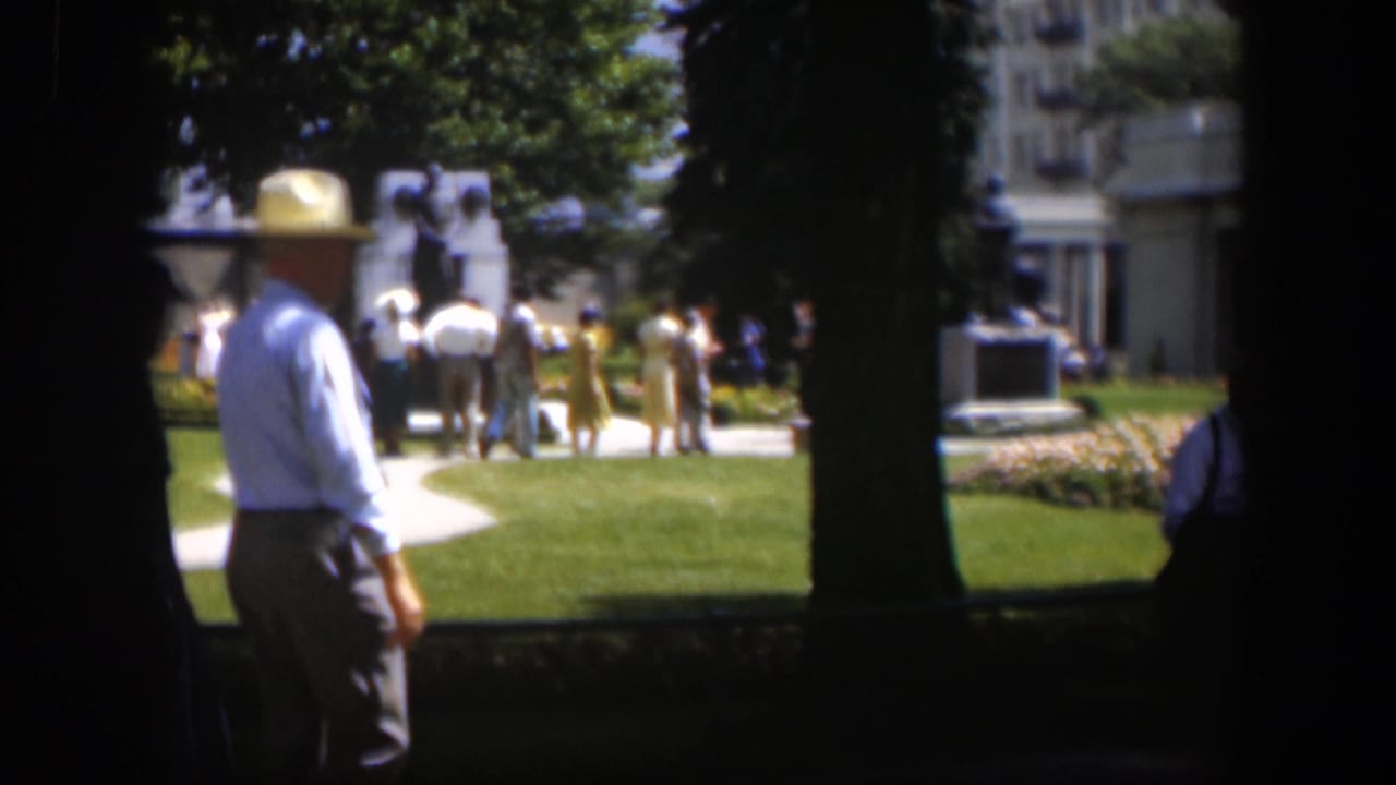 A man in a white hat stands near trees while visitors move near a monument and building.
