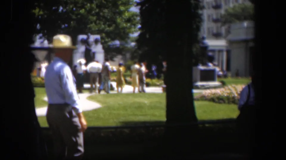 A man in a white hat appears in the foreground of a 1951 Wyoming park scene, with visitors, trees, a monument, and a building behind him.