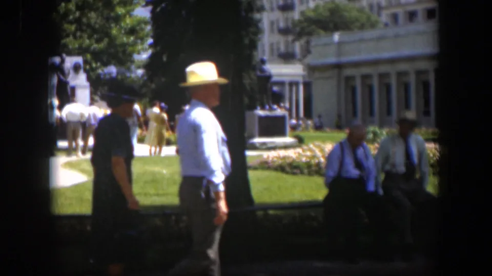 1951 Wyoming park frame showing adults near a path, including a man in a white hat, with flowers, a monument, and a building in the background.