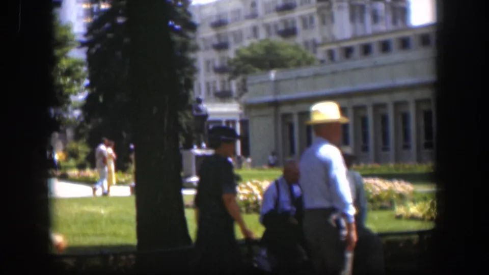 Adults in hats occupy the shaded foreground of a 1951 Wyoming park view, with lawn, flowerbeds, trees, and a building behind them.