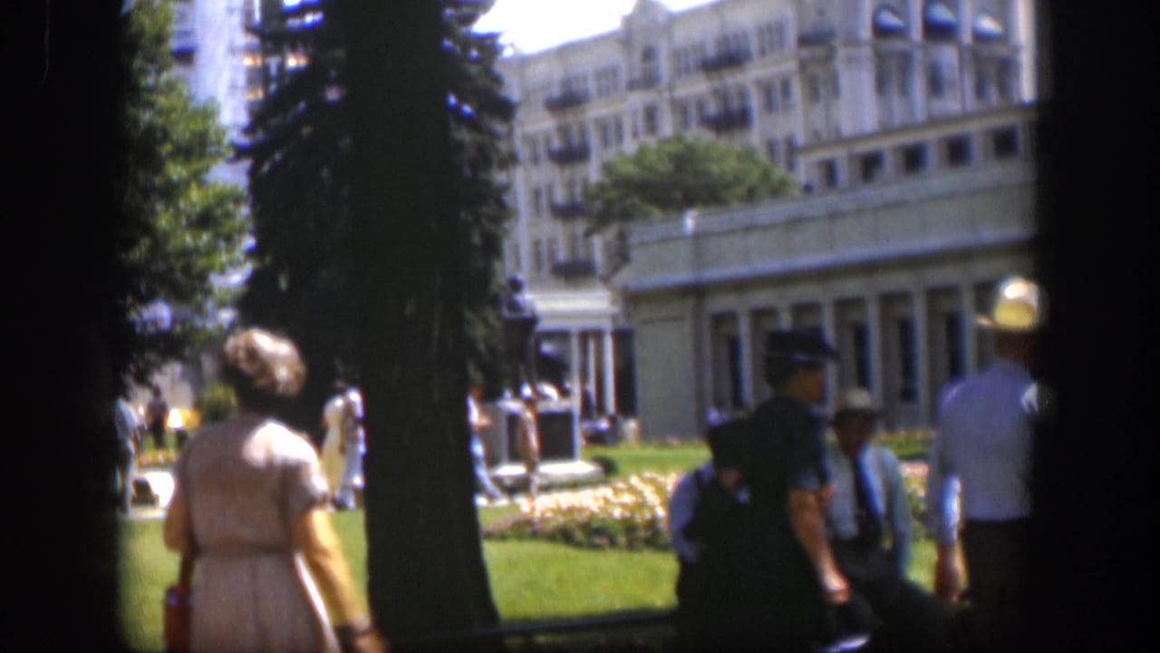 1951 Wyoming home-movie frame showing a woman near a lawn, trees, and a large public-looking building through dark foreground framing.