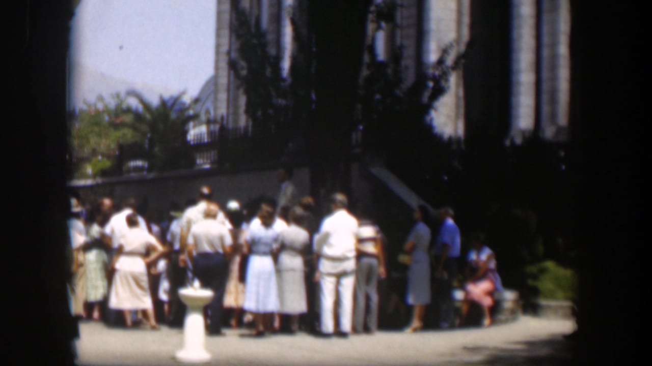 Group of adults gathers outside a large building beside a white pedestal and greenery.