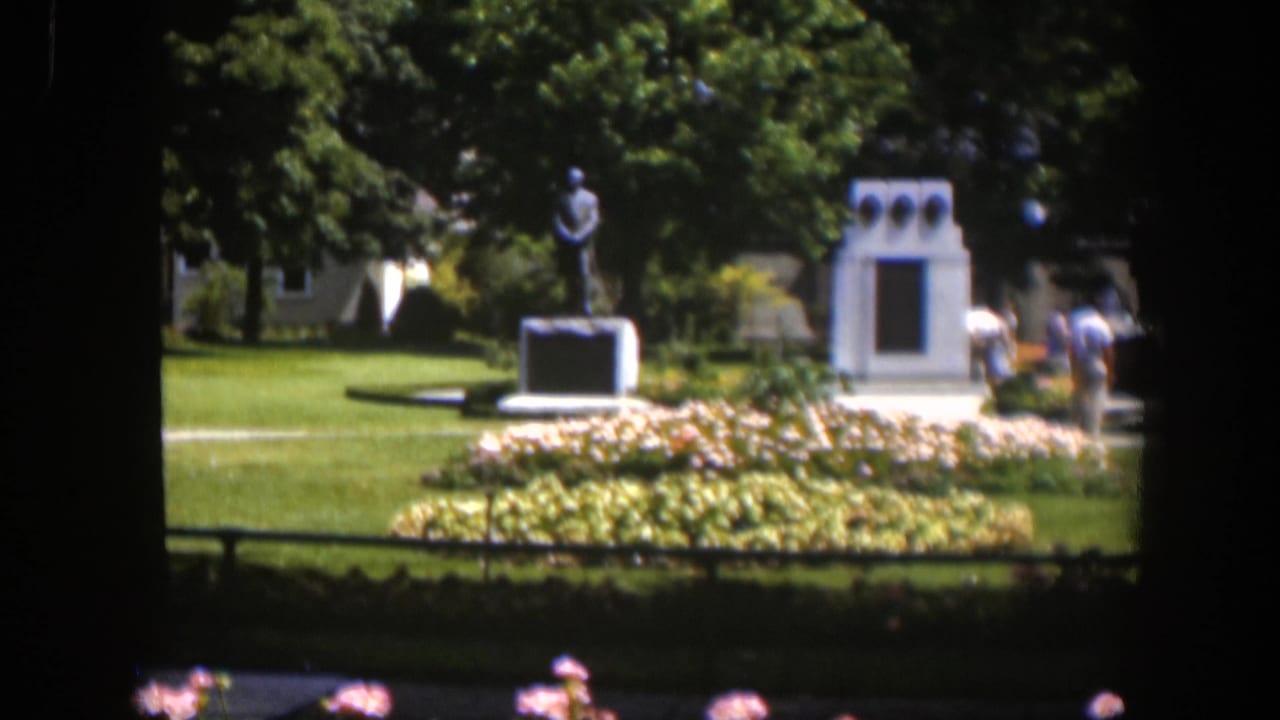 Visitors stand near a statue and white memorial structure in a sunny park.