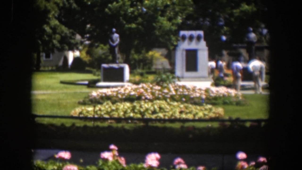 A distant 1951 Wyoming park view shows visitors moving around a statue and white memorial structure beside planted beds.