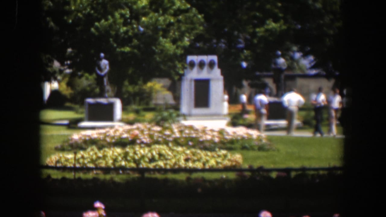 The frame shows a Wyoming park monument scene with visitors clustered near a pedestal, white structure, flower beds, and shaded trees.