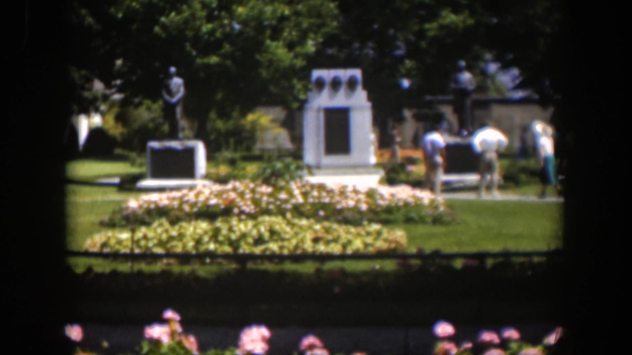 Distant home-movie view of a 1951 Wyoming park landmark with a statue, white memorial structure, flowers, grass, and visitors.