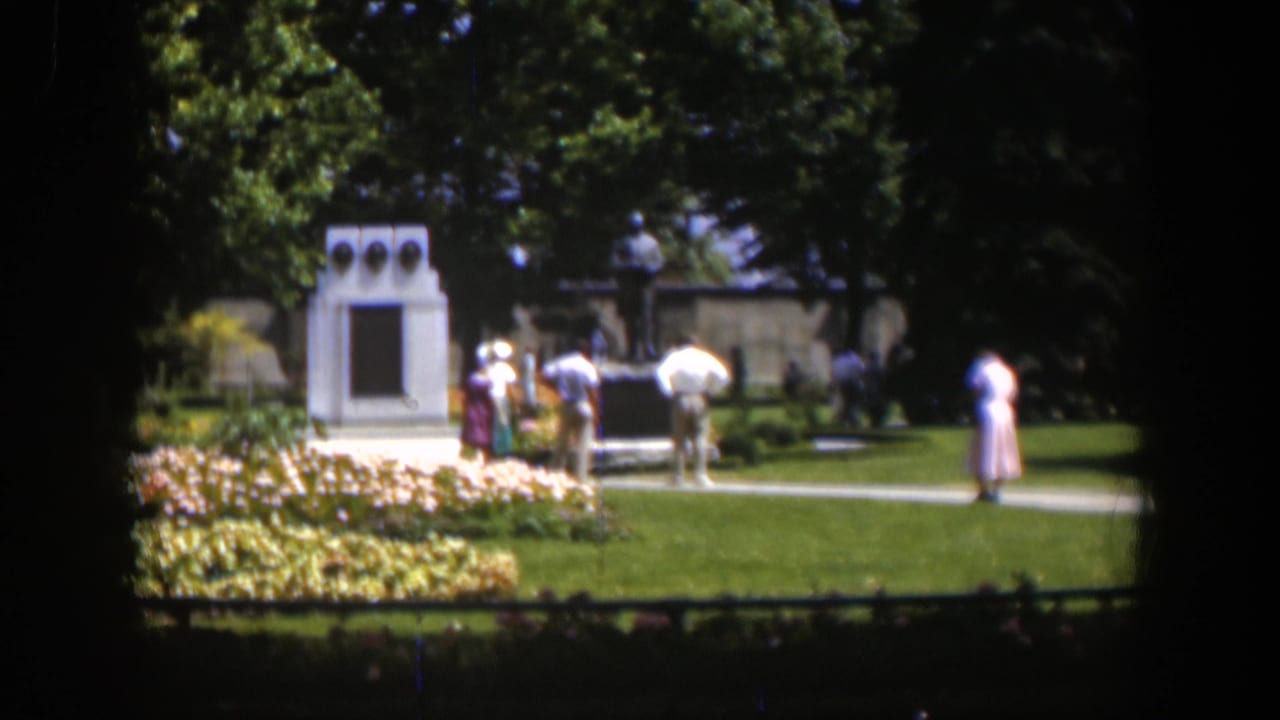 Adults walk and stand near a white monument and flowerbeds in a sunny park.