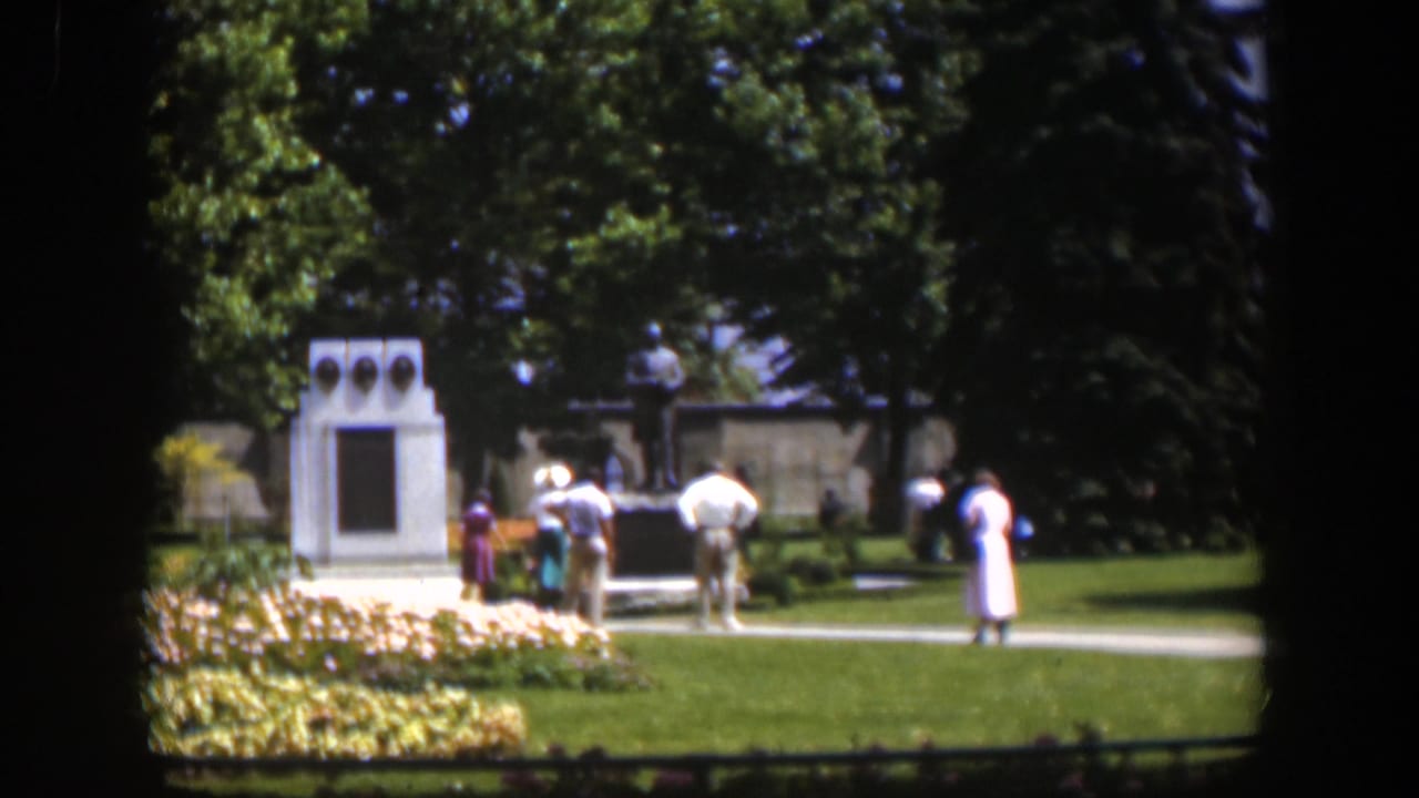 1951 Wyoming park scene with adults in period clothing near a statue, white monument, lawn, and trees.