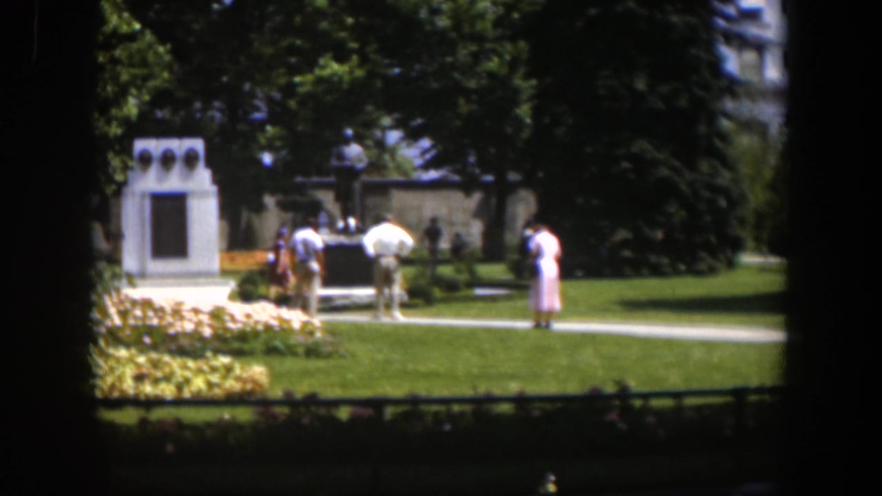 1951 Wyoming 8mm view of adults crossing a sunny park path near a white monument and flowerbeds.