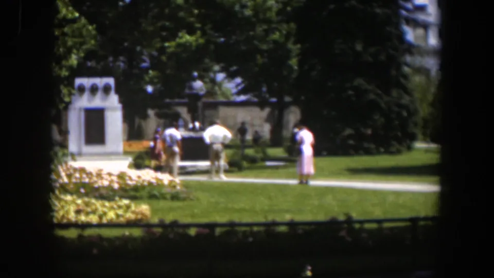 1951 Wyoming 8mm view of adults crossing a sunny park path near a white monument and flowerbeds.