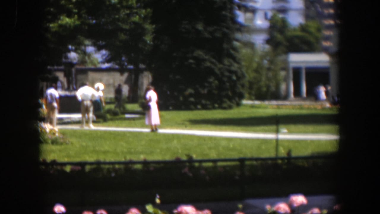 1951 Wyoming home-movie frame with visitors near a park statue, green lawn, trees, and a small structure in the distance.