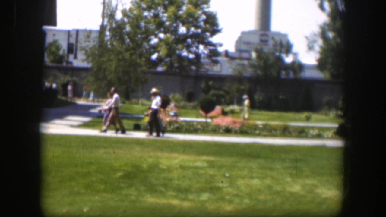 1951 Wyoming home-movie frame showing people on a park walkway, green lawn, flowerbeds, trees, and a large background building.