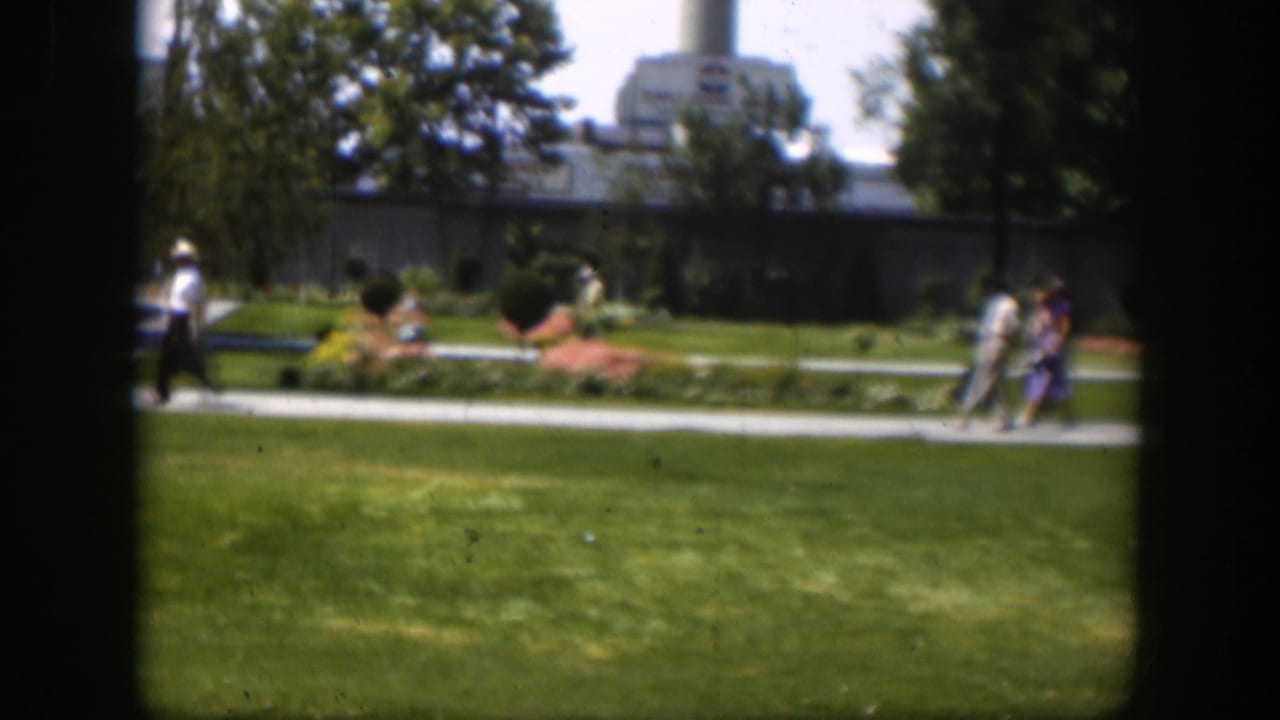 1951 Wyoming park scene with small groups of people walking past flowerbeds, lawns, trees, and an industrial-looking building.