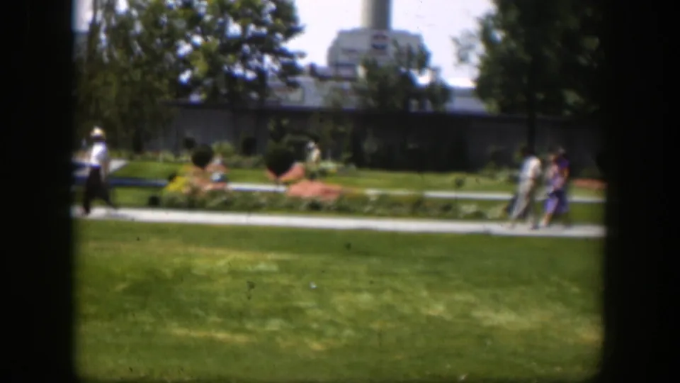 1951 Wyoming park scene with small groups of people walking past flowerbeds, lawns, trees, and an industrial-looking building.