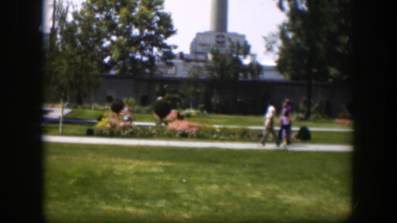 1951 Wyoming home-movie view of two pedestrians crossing a park path near flowerbeds, lawn, trees, and a distant large structure.