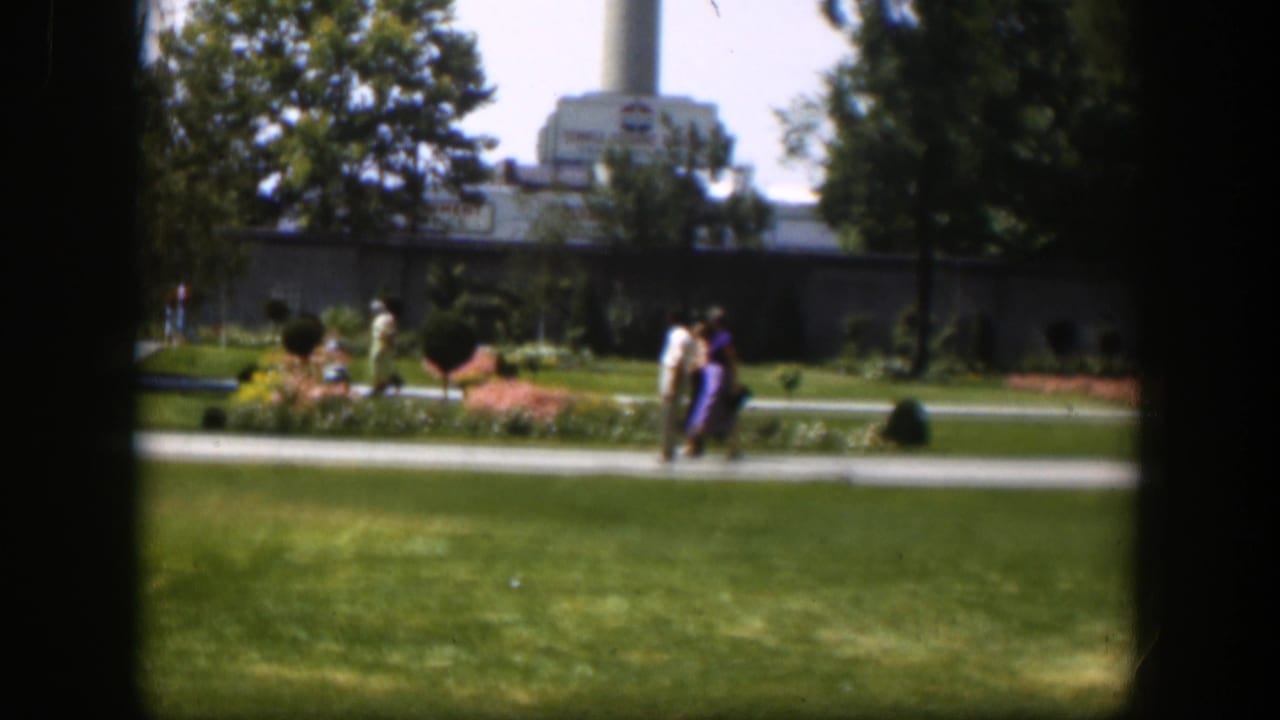 1951 Wyoming park frame showing pedestrians in period clothing near flowerbeds and lawns, with a large building and smokestack beyond.
