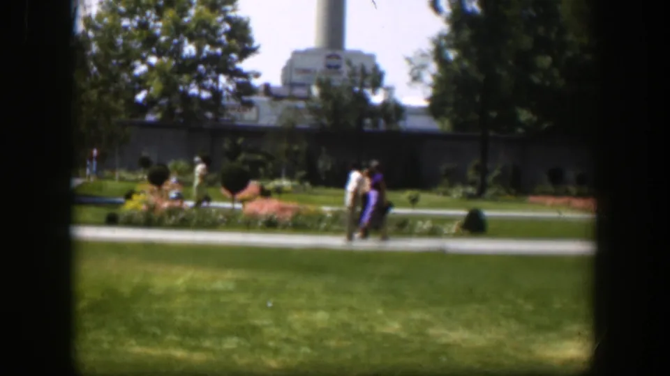 1951 Wyoming park frame showing pedestrians in period clothing near flowerbeds and lawns, with a large building and smokestack beyond.