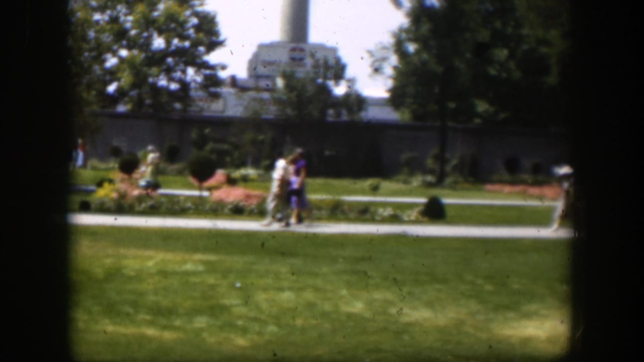 1951 Wyoming home-movie frame showing adults walking through a manicured park with a smokestack and industrial building in the background.