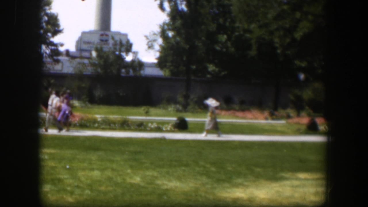 1951 Wyoming park scene with walkers on a paved path, lawns and flowerbeds in front of a distant industrial building and smokestack.