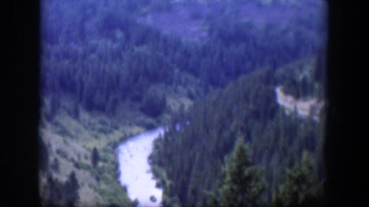 1951 Wyoming home-movie frame with a river bending through conifer-covered hills, seen through dark 8mm frame edges.