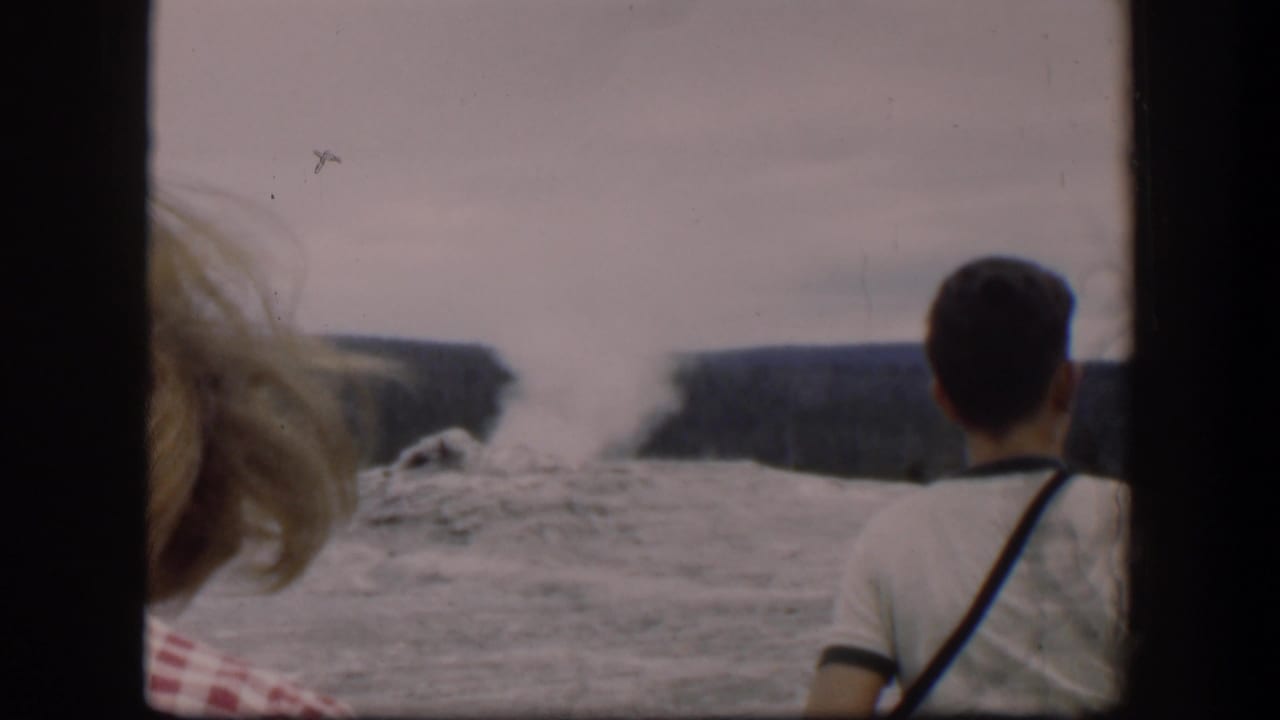 Spectators watch a steam plume rise from a rocky geyser area behind dark film-frame borders.