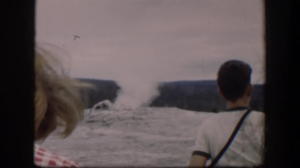 1951 Wyoming 8mm frame showing two spectators at the edge of view watching a geyser-like steam plume rise against distant ridges.