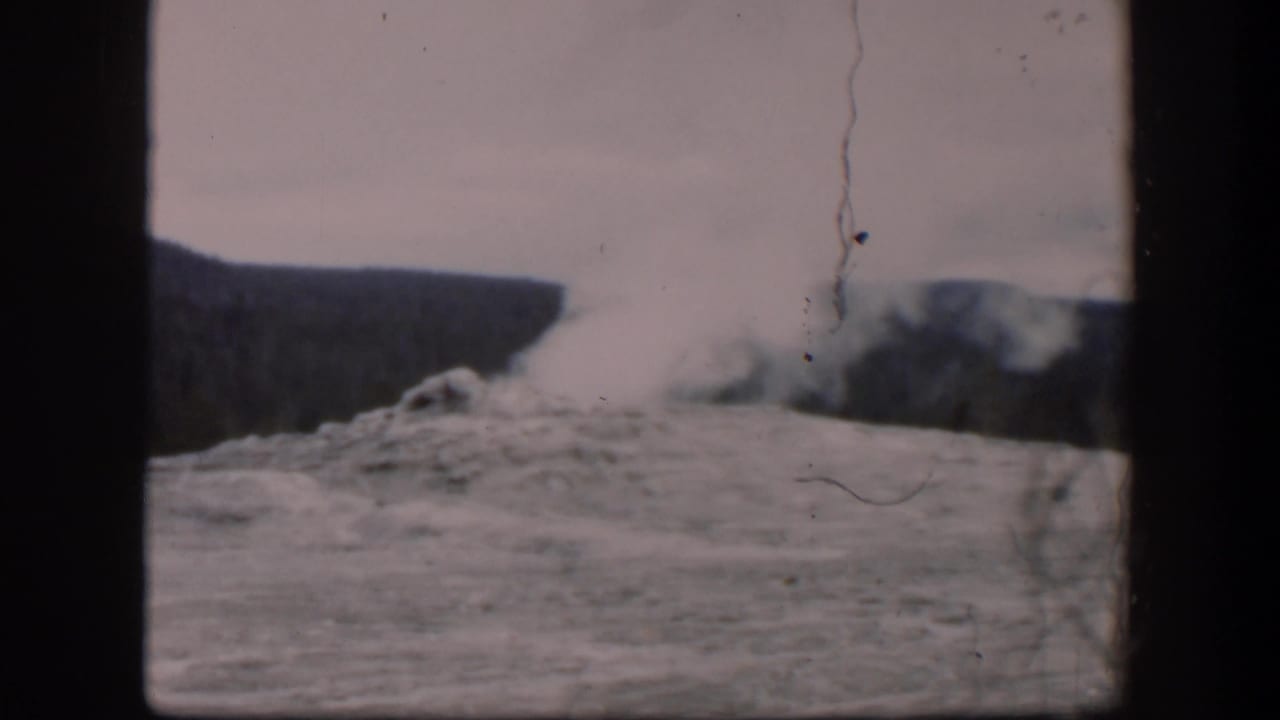 Steam and spray rise from a rocky geothermal area with ridges in the background.