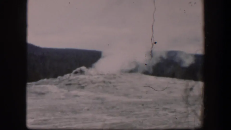 1951 Wyoming home-movie frame of a geyser-like eruption, with pale steam and spray rising above rocks and dark forested ridges.