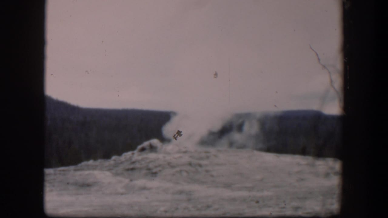 Geyser steam rises from rocky ground with dark ridges behind it.