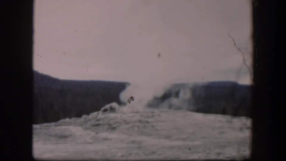 1951 Wyoming 8mm frame showing a geyser plume rising from a rocky vent with forested hills in the background.